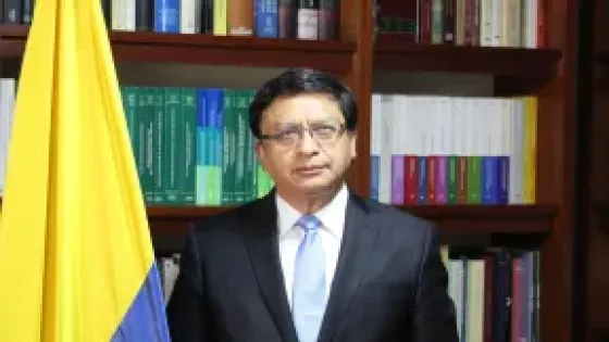 Fotografía de un magistrado de la Corte Constitucional de Colombia, de pie junto a la bandera nacional y frente a una biblioteca con textos jurídicos. La imagen acompaña la noticia sobre la ponencia que propone anular la reforma pensional por presuntas irregularidades en su trámite en la Cámara de Representantes.